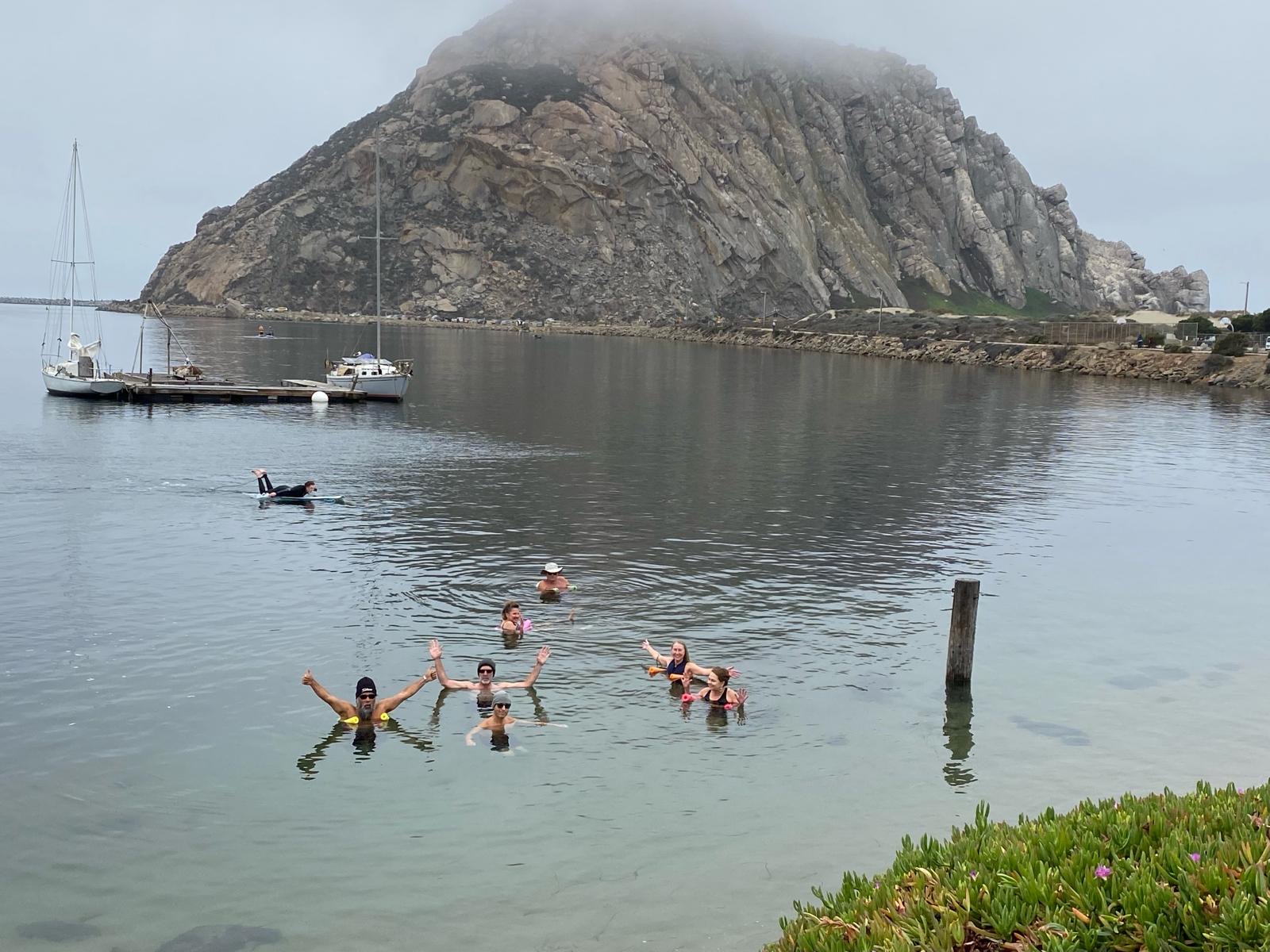 A group of odders waving and giving thumbs up from the water in front of Morro Rock