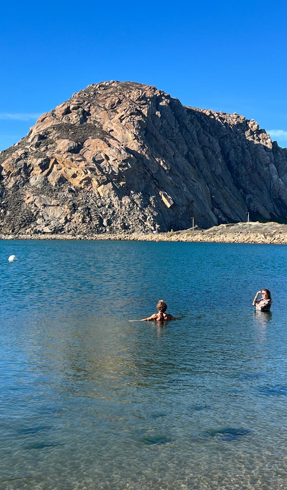 Two swimmers in deep blue water with Morro Rock towering behind them