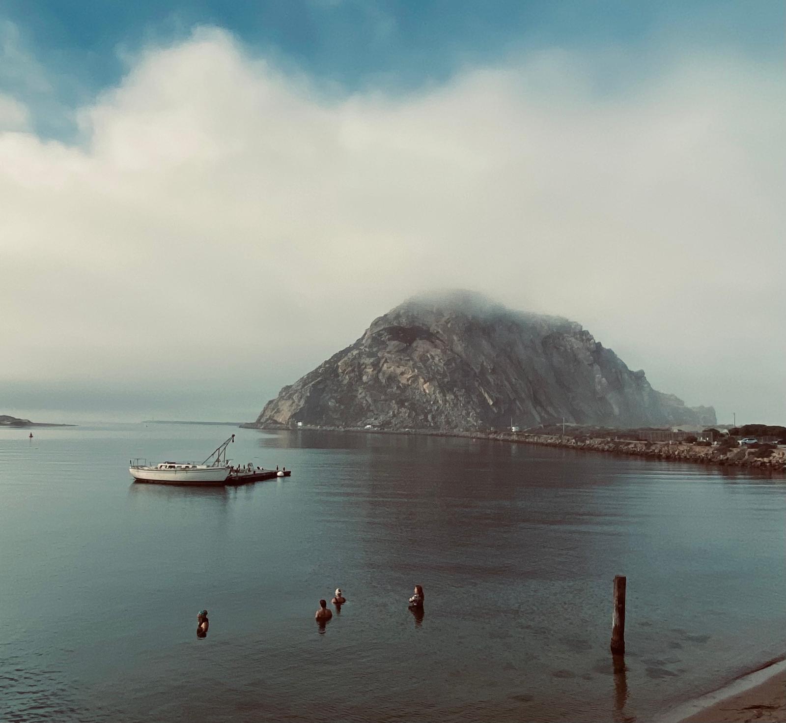 Morro Rock wrapped in low cloud with a small boat and swimmers in the foreground